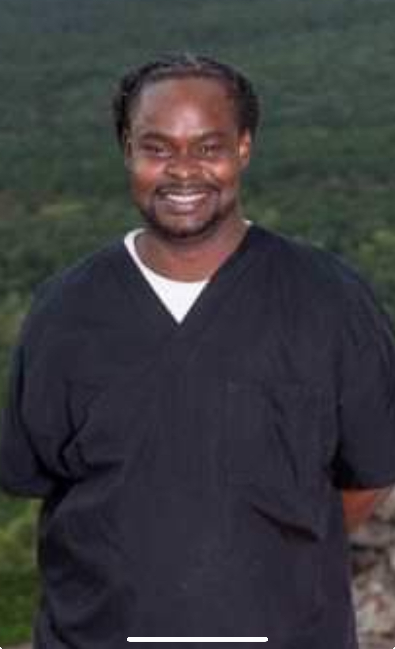 Smiling man in black scrubs with nature backdrop.